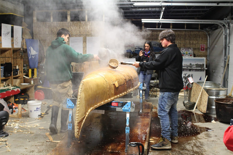 Niigaunii and Dawson pouring hot cedar water over the hull of the jiimaan to prepare for etching of the winter bark.